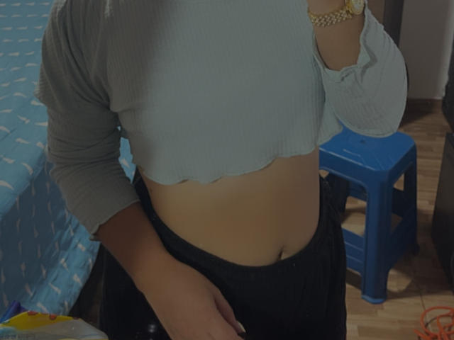 SarahGood's midriff shows a slight belly button peek, wearing a gray ribbed crop top and black pants, with a blue stool and wooden floor in the background.