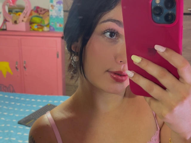 SarahGood taking a mirror selfie with pink lipstick, long nails, and pink bra straps, holding a pink phone, showing her face and upper chest. Her tits are small.