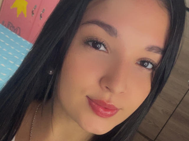 SarahGood has shiny black hair, blue eyes, and red lipstick. She's wearing pearl earrings and a gold necklace, with a cute smile. Her tits are not visible.