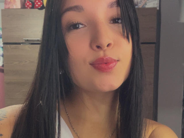 SarahGood has pouty pink lips, straight black hair, and a slight tan. She's wearing a white top with a necklace, and has a small tattoo on her right arm.