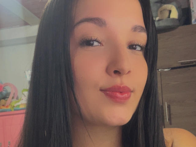 SarahGood with straight black hair, brown eyes, and glossy pink lips, giving a slight pouty smile. Her light skin has a subtle pinkish tone. She's looking to the side.