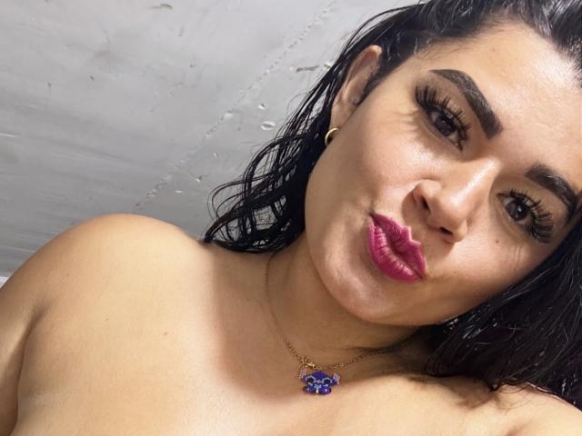Antonella Bellas tem cabelos pretos e úmidos, sobrancelhas escuras, cílios longos, batom rosa e um colar de ouro com uma flor azul. Ela está fazendo beicinho com seus lábios brilhantes e olhando para a câmera..