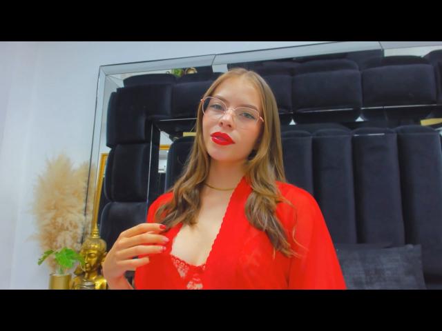 AnnaFer con rossetto rosso, occhiali e top rosso trasparente, che mostra i suoi seni con un reggiseno in pizzo, una mano sul petto, capelli biondi, e un poggiatesta nero sullo sfondo..