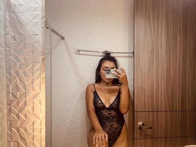 AmeliRoux portant une combinaison noire en dentelle avec un décolleté en V profond, laissant entrevoir ses seins et ses parties intimes à travers le tissu transparent, se prend en photo devant un miroir dans une pièce en bois..