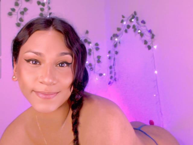 AndreaFeoxx with dark braided hair, thick eyebrows, and winged eyeliner, smiling at the camera, topless, showing her bare back and blue thong, pink lighting in the background.