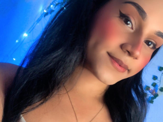 AndreaFeoxx has fair skin, black hair, and a slight smile with pink blush on her cheeks, wearing eyeliner, and a thin necklace, with blue-lit background.