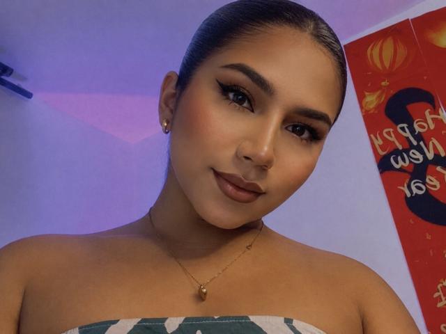 AndreaFeoxx with smooth brown skin, dark eyes, and full lips, wearing a green and white strapless top, gold necklace, and earrings, looking at the camera with a slight smile.