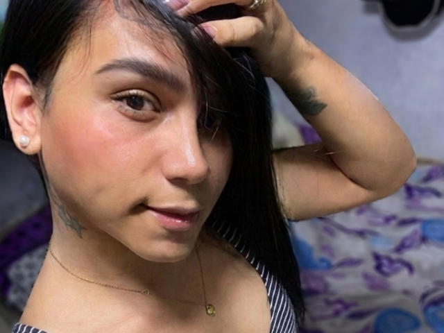 AndreaFeoxx with black hair, hand in hair, slight smile, tan skin, small tattoos, gold necklace, striped top, ear piercings, subtle makeup, bedroom setting.