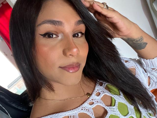 AndreaFeoxx with glossy pink lips, black eyeliner wings, brown eyes, tan skin, black straight hair, white crochet top, green pattern, hand touching hair, tattoo on right arm.