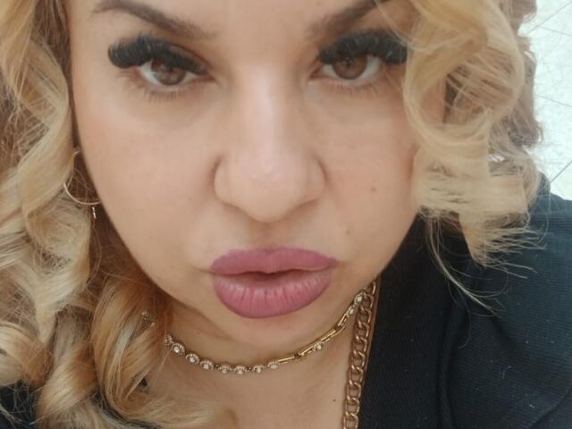 BelleInna has blonde curly hair, thick black eyelashes, pink lipstick, and a gold chain necklace. She's looking directly at the camera with a serious expression.