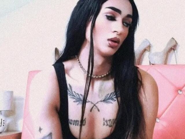 IrisIrs with long black hair, heavy eyeliner, and dark lipstick, wearing a black tank top, showing tattoos across her chest and arms, and a pearl necklace.