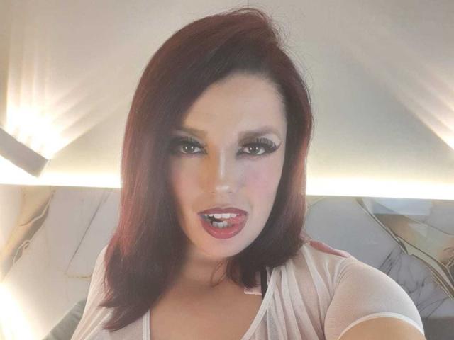 SachyMoon met rood haar, make-up en een wit shirt, die haar roze lippenstift laat zien en haar mond is lichtjes open, kijkend recht in de camera. Haar borsten zijn nauwelijks zichtbaar..
