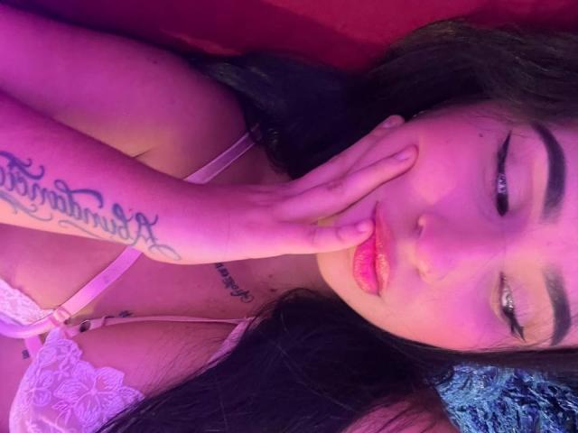 Hannah Becker, con biancheria intima rosa, labbra piene e un tatuaggio sulla mano che le copre la bocca, ha gli occhi azzurri, i capelli scuri ed è sdraiata in un ambiente illuminato di viola e rosso..