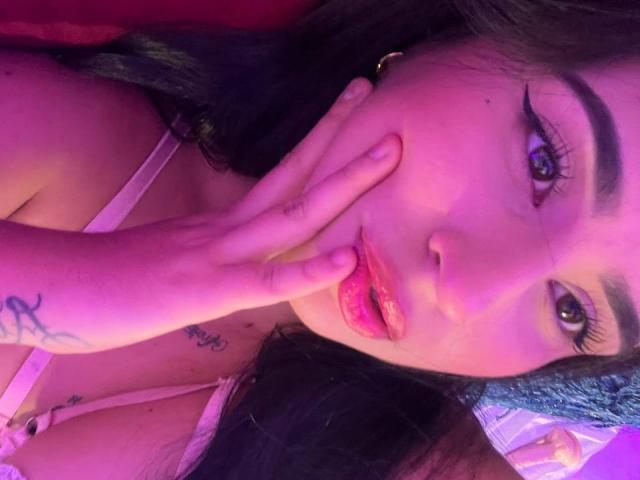 Het gezicht van HannahBecker, paars-roze verlicht, met een hand die de mond bedekt met één vinger, donker haar, eyeliner, lipgloss, kleine tatoeages op de hand en nek, zichtbare decolleté in een roze beha..