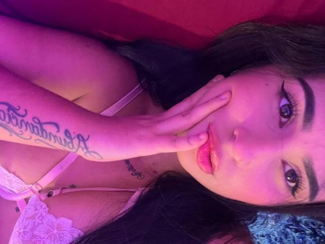 HannahBecker with pink lipstick, brown eyes, and long dark hair, touching her face, showing a tattoo on her left hand, wearing a white lace bra, under purple lighting.