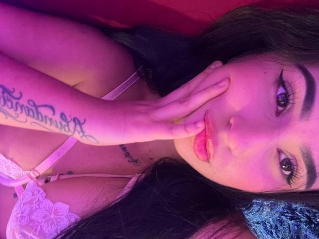 Hannah Becker, avec du rouge à lèvres rose, des cheveux foncés et un tatouage sur la main, se cachant la bouche, portant un soutien-gorge en dentelle blanche, avec un éclairage violet et rose, regardant la caméra, yeux roses..