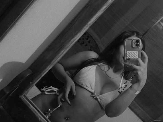 AntonellaMonroe in a black-and-white selfie wearing a white, crocheted bikini top, showing her tits and a belly button piercing, holding a phone with a textured case.
