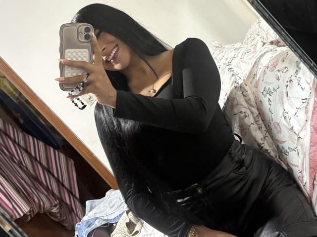 Antonella Monroe, avec de longs cheveux noirs, souriante, portant un haut noir à manches longues et un pantalon noir, tenant un téléphone dans une photo de profil prise dans un miroir, montrant ses seins et ses fesses..