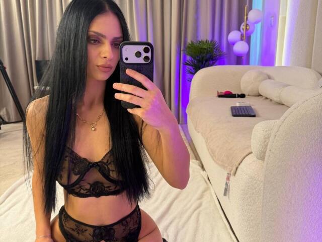 IliriaKara with long black hair, wearing black lace bra and panties, taking a mirror selfie, showing her tits and ass, holding a phone with a gold necklace.