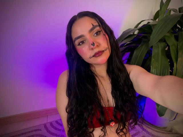 FenixAlthea has long black wavy hair, dark makeup, red lipstick, nose ring, and black lipstick scars. She's wearing a red bra, with purple and blue lighting.