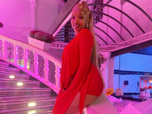 CaramelElla with long blonde hair, red long-sleeve dress, and white thigh-highs, smiling, standing on stairs with purple and pink lighting, looking over her shoulder.