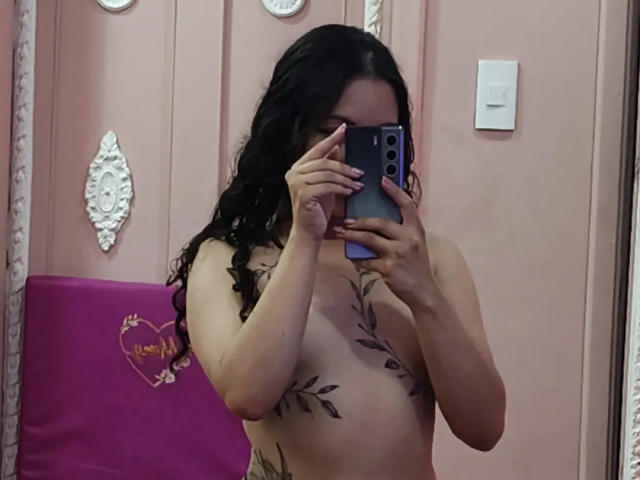 MiiaBennet with dark wavy hair, holding a phone, covering her face, showing tattoos on her chest and holding a purple bag, standing topless.