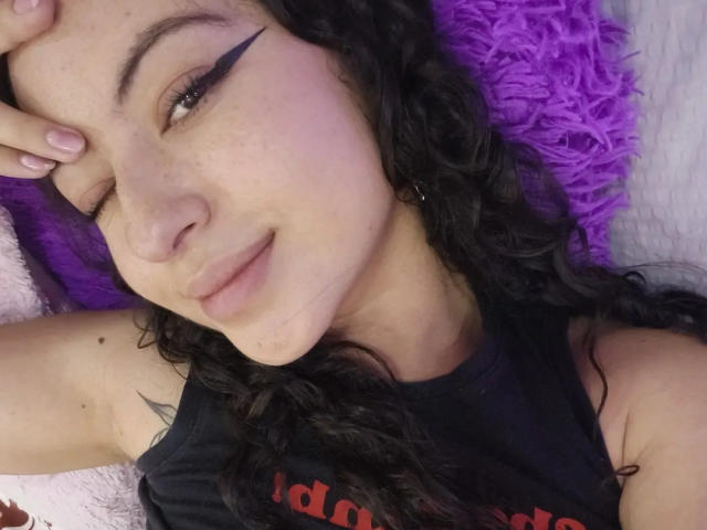 MiiaBennet, liggend op een paars kussen, knipoogt met zwarte eyeliner, heeft krullend zwart haar, bleke huid, roze lippen en draagt een zwart topje zonder mouwen. Haar hand rust op haar voorhoofd..