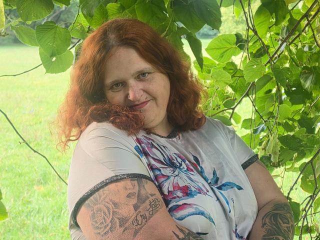 Hotsunny20-sex has red curly hair, tattoos on her arm, and is smiling under green leaves. She's wearing a white shirt with blue and red floral patterns.