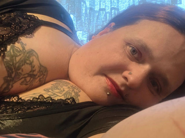 Hotsunny20-sex with tattoos on her shoulders and chest, black lace bra, nose and lip piercings, lying on a black leather surface, blue curtain in background.