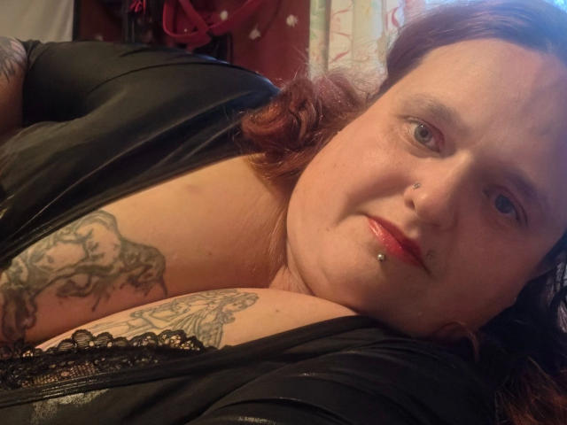 Hotsunny20-sex, with red hair, nose ring, and lip piercing, lies down, showing tattoos on her tits and chest, wearing a black top, in a dimly lit room.