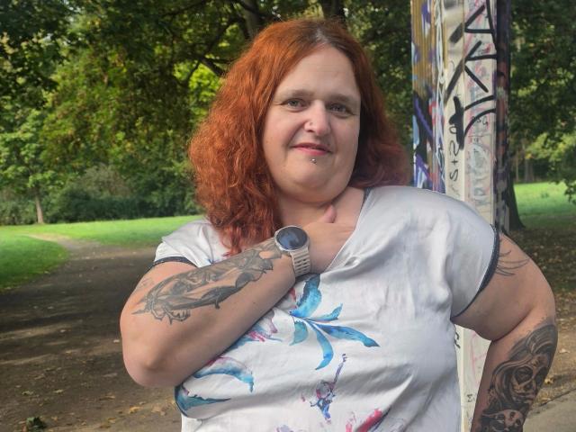 Hotsunny20-sex with red curly hair, white t-shirt, tattoos on arms, watch on wrist, piercing on chin, standing by graffiti-covered pole, green park in background, smiling slightly.