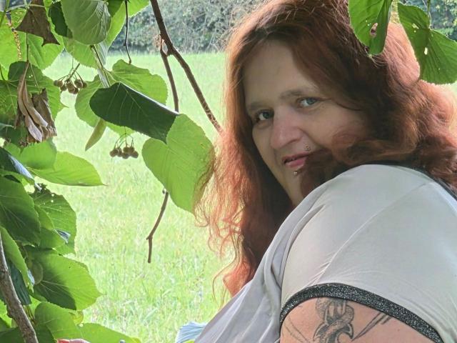 Hotsunny20-sex with red hair, small nose ring, white shirt, visible arm tattoo, smiling, leafy green background, grassy field, sunlit, looking back over shoulder, natural light.