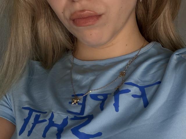 O rosto de SugarrX, com lábios carnudos, vestindo uma camiseta azul e um colar de prata com um pingente em forma de estrela, mostrando um pouco de seu cabelo loiro..