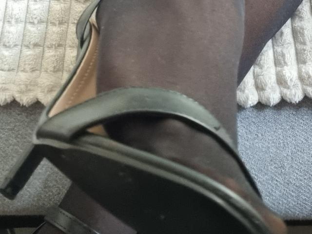 SexyMirra's dark brown leg with green high-heeled sandal, showing the sole and strap, resting on a textured carpet. The sole is slightly worn, and the strap is visible.