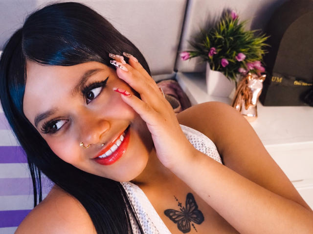 Nahara, une femme aux cheveux foncés, au rouge à lèvres rouge, et avec un tatouage de papillon sur sa poitrine, souriante, touchant son visage, montrant ses longs ongles, et portant un haut en dentelle blanche..