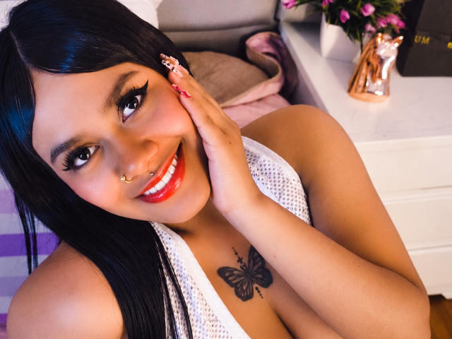 NaharaFarfalle with dark hair, red lipstick, and white mesh top, smiling, showing her butterfly tattoo, touching her cheek, nose ring, and long nails, in a bedroom setting.