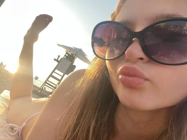 ShanteWellendorf with pouty lips, wearing big purple sunglasses, lying on beach, legs up, sun shining, brown hair, pink bikini, and a slight tan, showing her ass in the background.