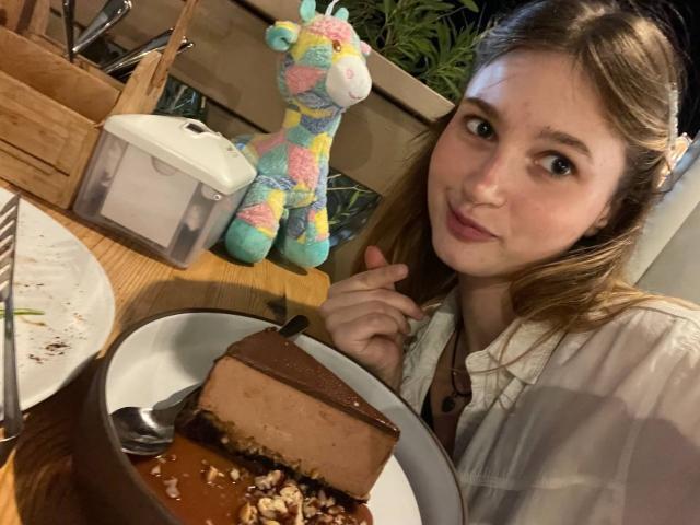 ShanteWellendorf with brown hair, white shirt, and light makeup, holding fork, smiling at camera. Chocolate slice with nuts on plate, colorful stuffed toy, wooden table, and sugar jar.