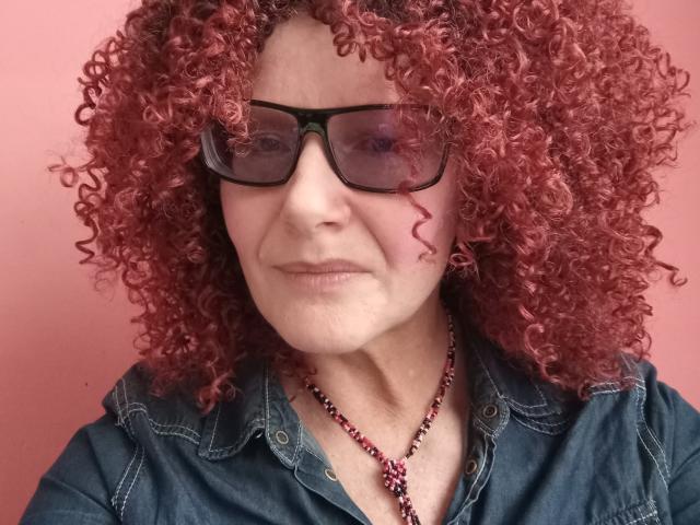 ChristineSweety with curly red hair, black glasses, and a denim jacket, wearing a beaded necklace, looking slightly annoyed. Her tits are small, and her ass is not visible.