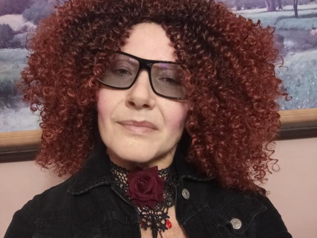 ChristineSweety, con cabello rojo rizado, gafas negras, un collar de encaje negro con una rosa roja, y una camisa de mezclilla negra, sonriendo ligeramente. Sus senos están cubiertos por la camisa..