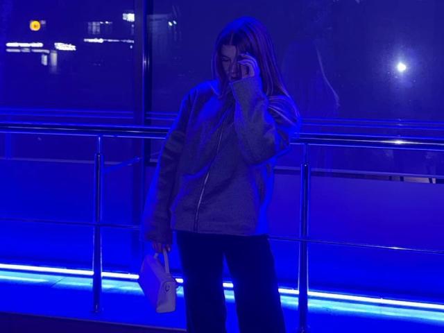 NorahHabrock stands in blue-lit indoor space, wearing a grey jacket, black pants, and holding a white purse while talking on a phone, with a reflective metal railing.