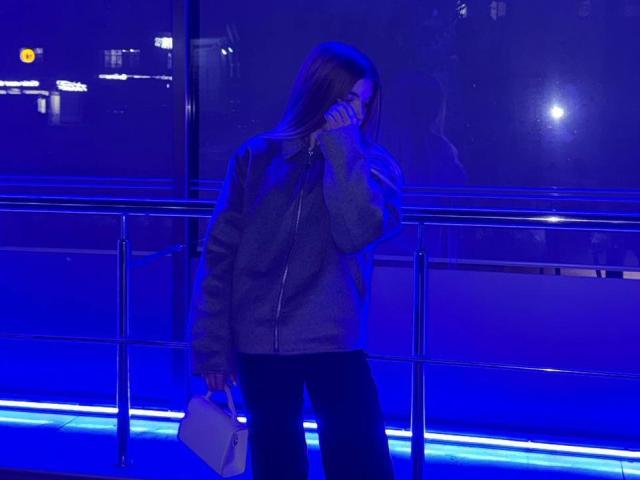Norah Habrock, con lunghi capelli castano tendenti al blu, indossa una giacca grigia con zip e pantaloni neri, tiene una borsa bianca, si copre la bocca e si trova accanto a una ringhiera di metallo..