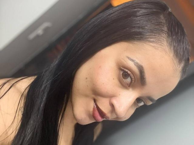 HikariChloe ha lunghi capelli neri, occhi marrone chiaro e sta sorridendo leggermente, con un rossetto rosso scuro. Ha piccole lentiggini sulla guancia e sta guardando a destra..