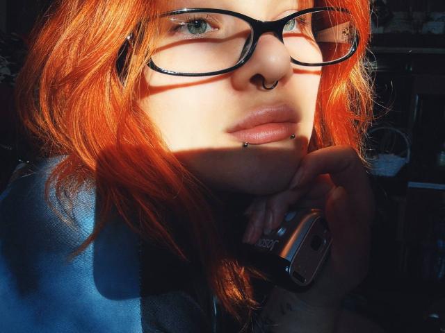 ClumsyNana with red hair, black glasses, nose ring, and lip piercing, holding a camera, sun casting shadow on her face, blue shirt, sunlight highlighting her features.