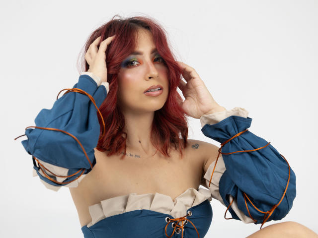MiaaRouge with red hair, wearing a blue off-shoulder dress with ruffled sleeves, touching her head, showcasing her tits and small tattoos on her chest.