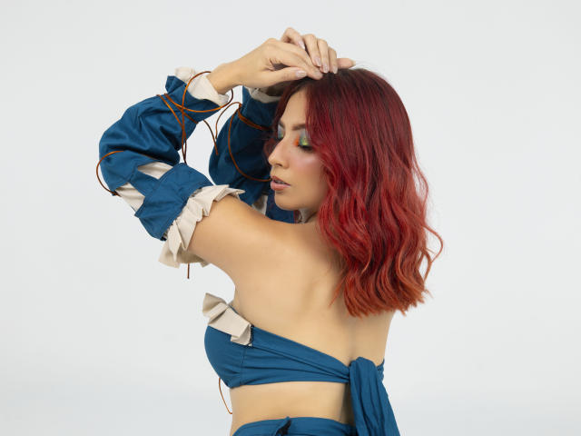 MiaaRouge con capelli rossi, top blu con spalline scoperte e polsini bianchi, braccia alzate, che mostra la schiena e il seno, trucco verde per gli occhi, capelli ondulati, sfondo bianco..