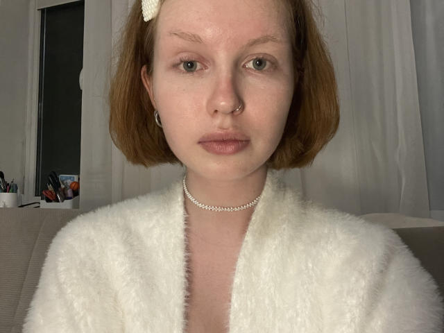 Deedra Domann tem cabelo curto e ruivo, uma faixa branca na cabeça, um piercing no nariz e um colar. Ela veste um roupão branco e fofo, tem pele clara e olhos verdes, e parece séria..