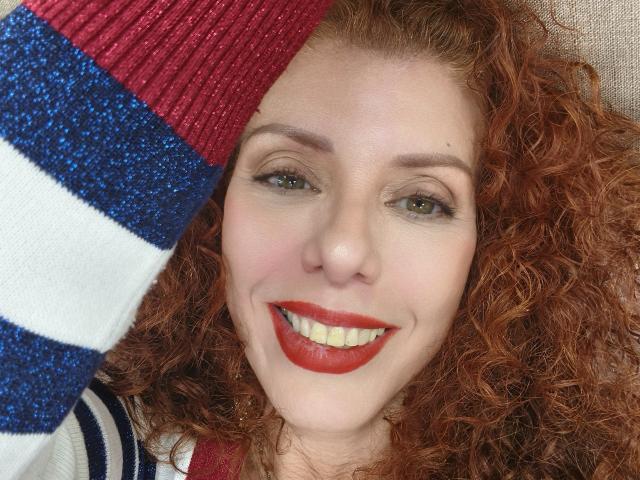 Khharha ha i capelli rossi ricci, gli occhi verdi e il rossetto rosso. Sta sorridendo e indossa un cappello a maglia blu, bianco e rosso che le copre parte della testa. Il seno non è visibile..