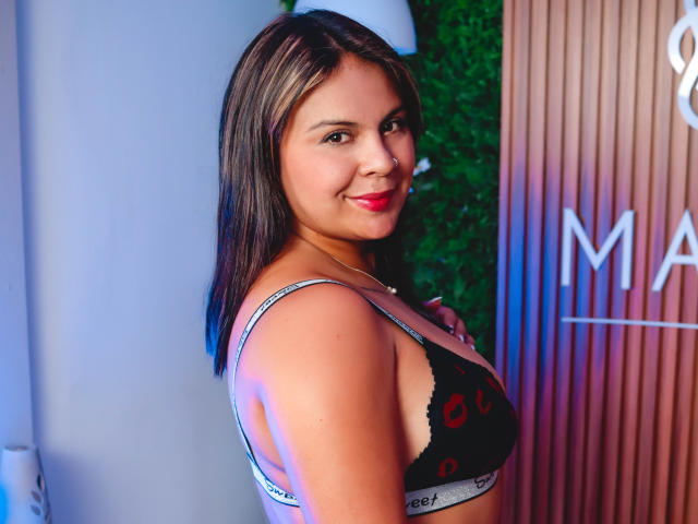 CamillaMoretti with medium brown skin, dark brown hair, red lipstick, smiling, wearing a black Calvin Klein bralette, showing her tits, standing against a wall with green and wood.