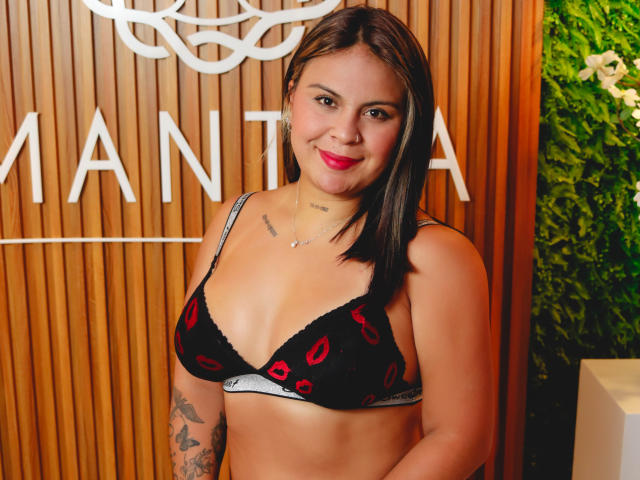 CamillaMoretti with medium tits, black bra with red lips pattern, tattooed arm, brown hair, red lipstick, wooden background, green plant, smiling, looking at camera.
